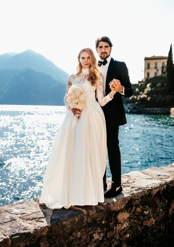 Brautpaar am Seeufer, die Braut trägt ein langärmliges Brautkleid mit weitem Rock und hält einen Brautstrauß