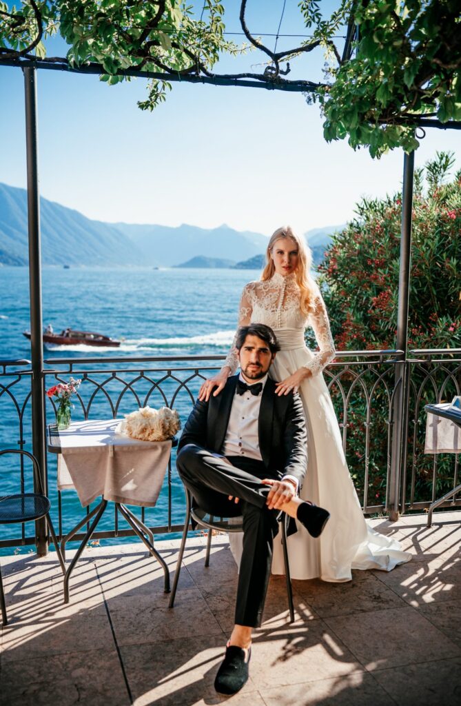 Braut im Spitzenbrautkleid steht auf einer Terrasse am See, der Bräutigam sitzt davor auf einem Stuhl